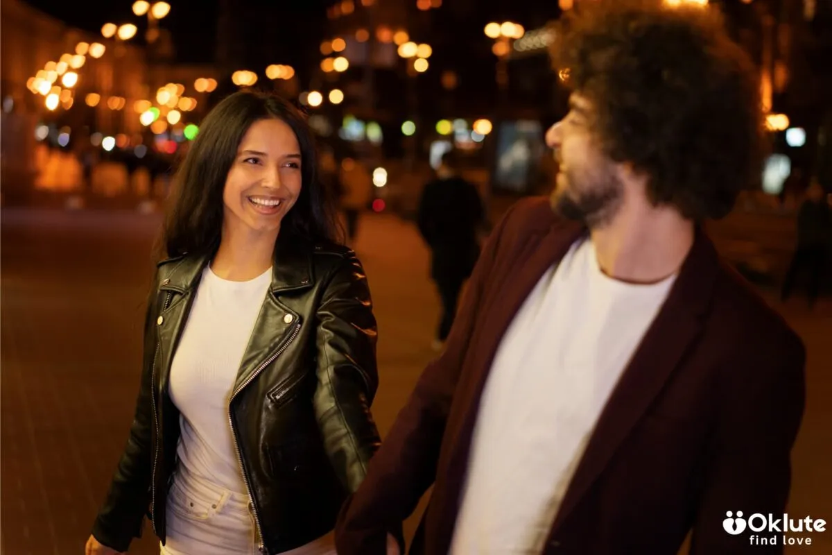 Um casal jovem, mulher com jaqueta de couro e homem de blazer, caminhando sorrindo em uma rua iluminada à noite.
