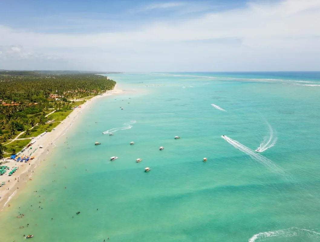 Praia com areia clara, mar azul e muitos barcos, ao fundo, vegetação e gente se divertindo.