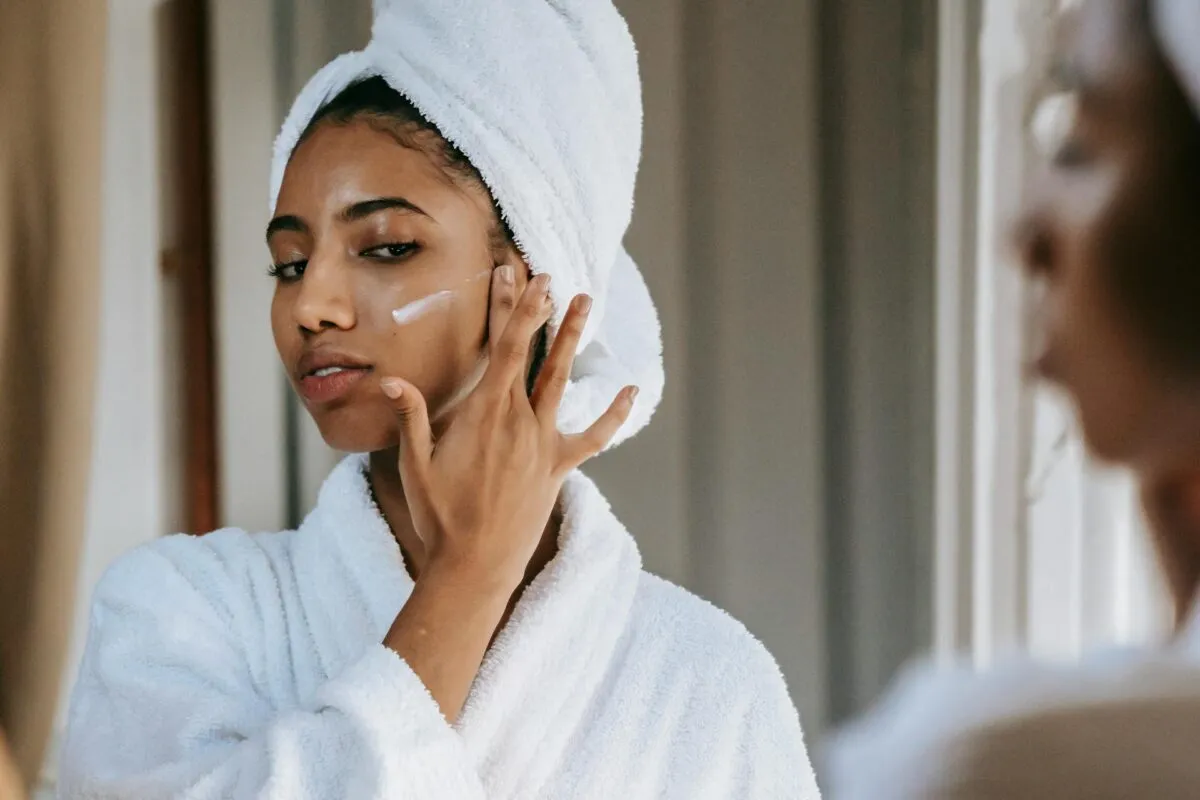 Mulher com toalha no cabelo aplica creme no rosto com expressão focada