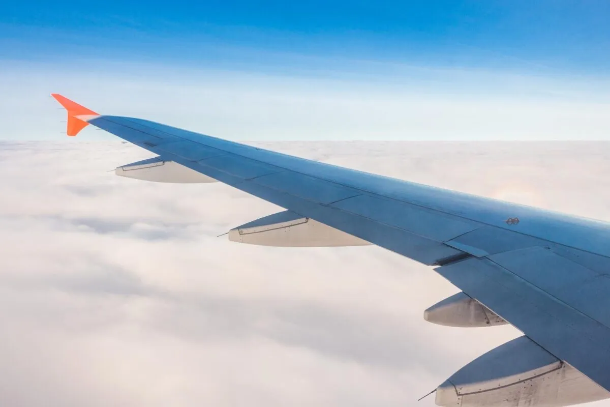 Asa de avião vista pela janela durante o voo, sobrevoando um tapete de nuvens brancas sob céu azul claro