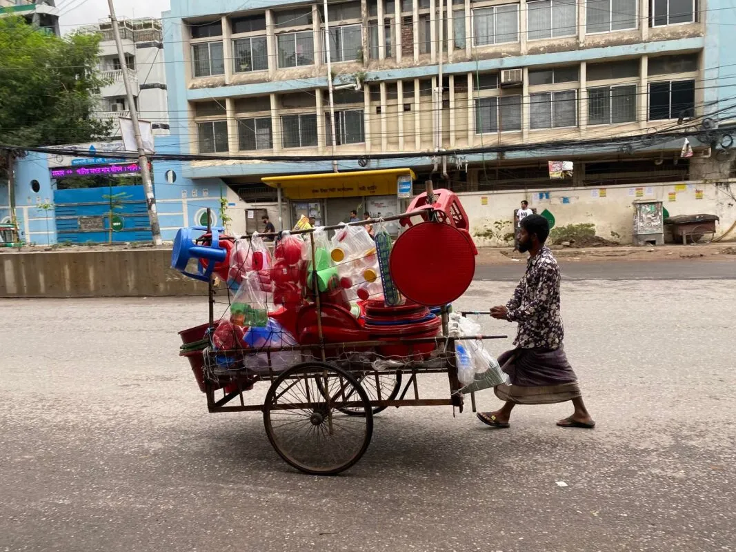Homem empurra carrinho com utensílios plásticos coloridos por uma rua movimentada de Dhaka, em Bangladesh