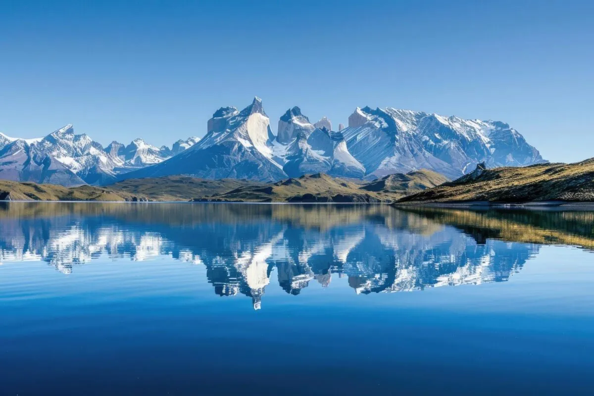 Picos nevados do maciço Torres del Paine refletidos nas águas calmas de um lago, sob céu azul, na Patagônia chilena