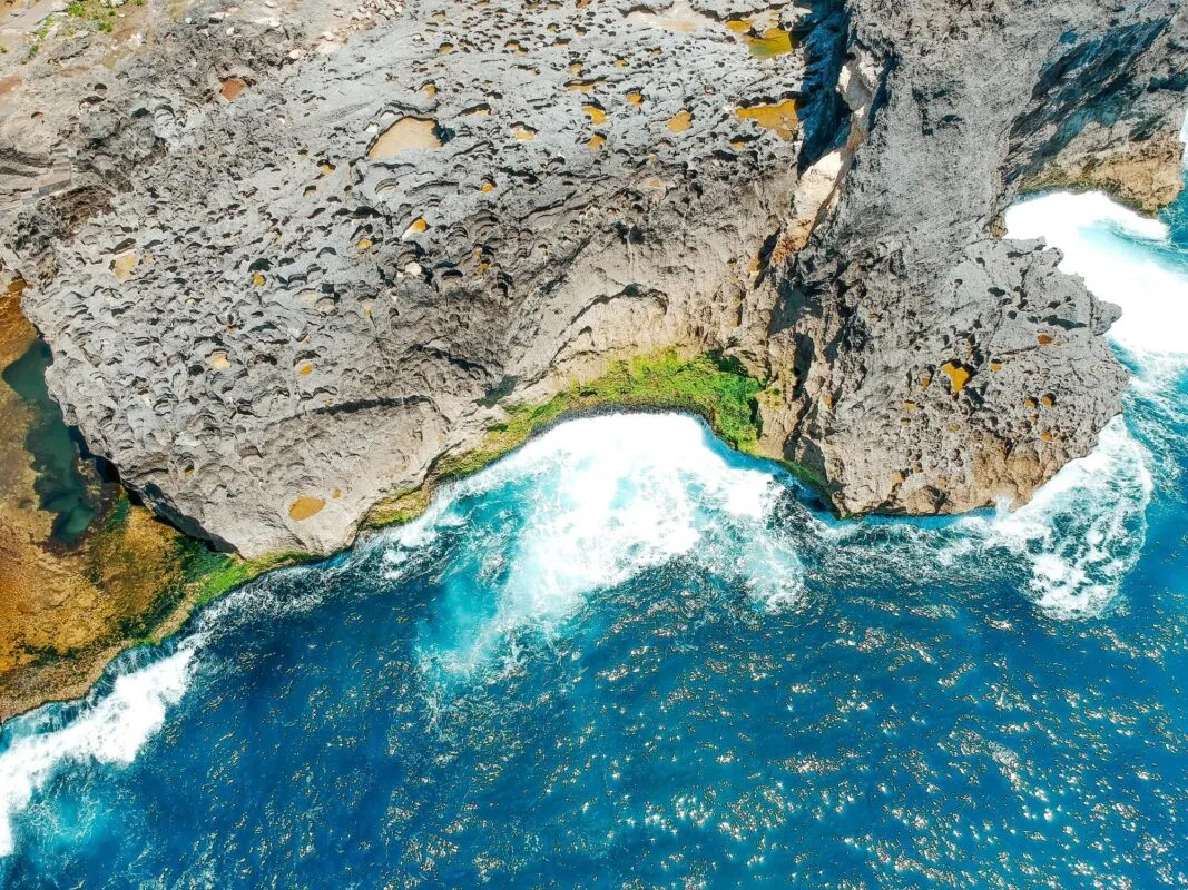 Vista aérea de costa rochosa vulcânica com ondas brancas quebrando contra o paredão escuro e mar azul intenso
