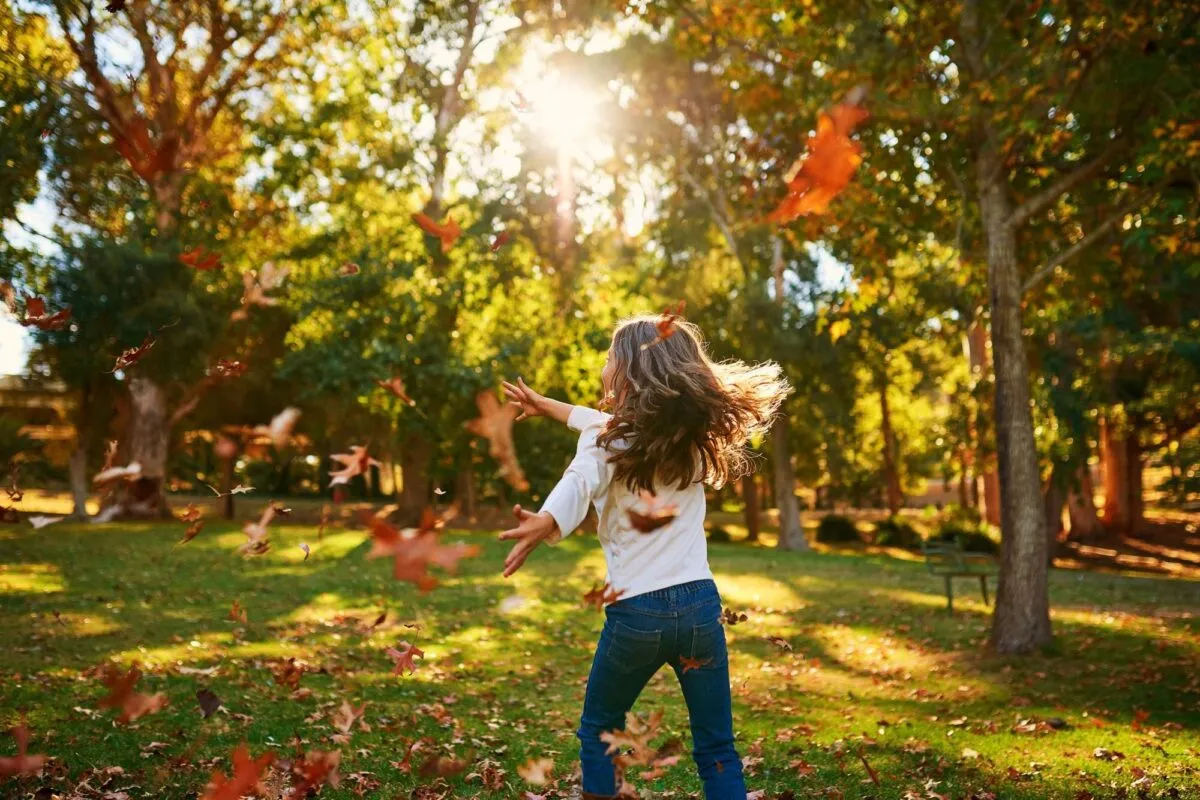Menina de costas brincando e jogando folhas de outono ao ar livre, em um parque com luz do sol entre as árvores