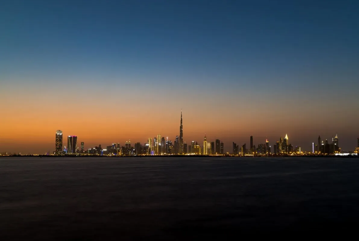 Vista do horizonte de Dubai ao entardecer, com arranha-céus iluminados e o Burj Khalifa ao centro, refletidos sobre a água