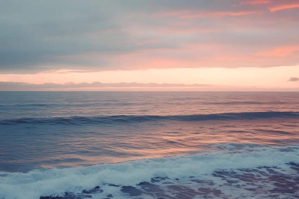 Mar calmo ao entardecer, com ondas suaves quebrando perto da praia e o céu em tons de rosa e cinza