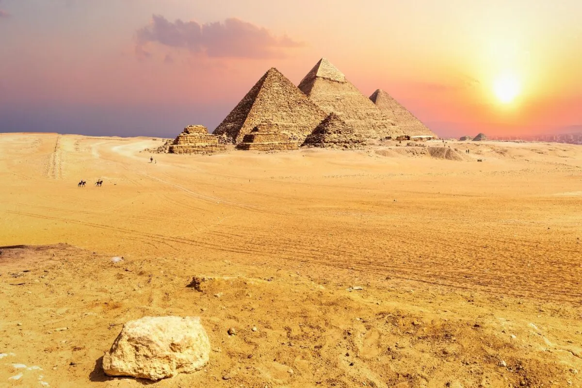 Pirâmides de Gizé iluminadas pelo pôr do sol, com o deserto em primeiro plano e o céu alaranjado ao fundo