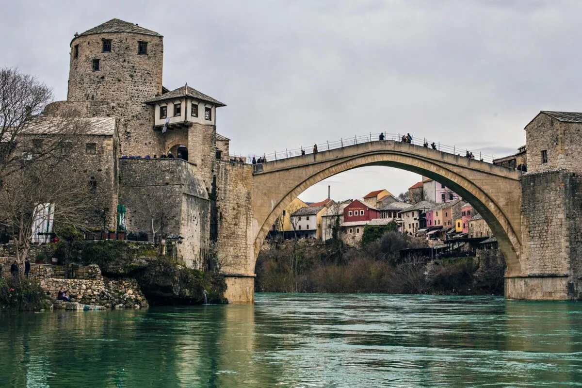 Ponte de pedra em arco sobre rio de águas esverdeadas, com casas antigas e torres de pedra ao fundo, na cidade de Mostar