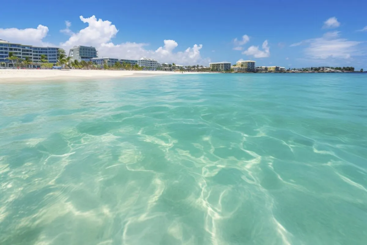 Praia de areia clara com água turquesa cristalina e prédios de resort ao fundo, sob céu azul com nuvens