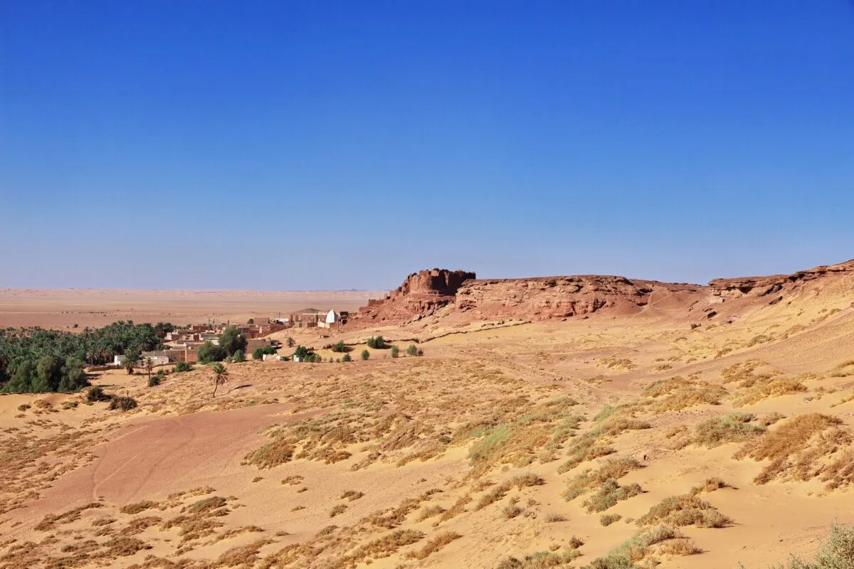 Antiga fortaleza de pedra avermelhada e oásis com palmeiras em meio às dunas do deserto do Saara, na Argélia, sob céu azul.