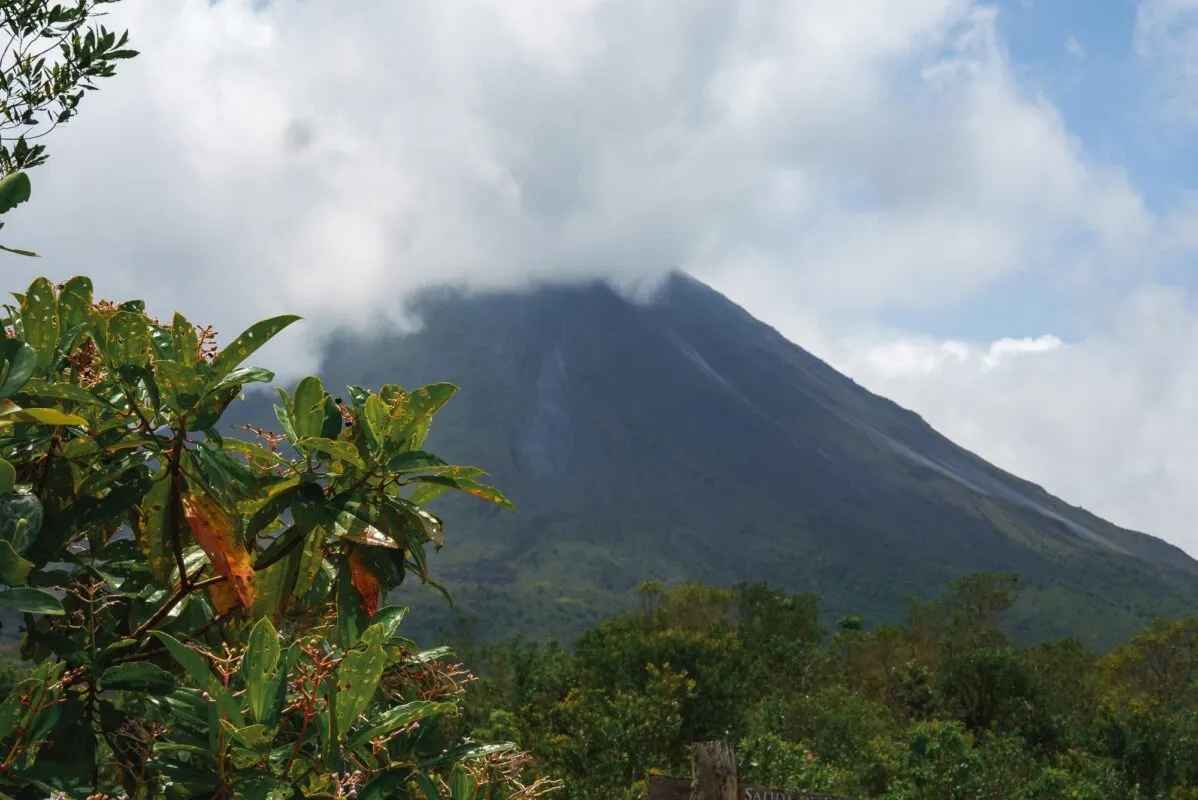 Vulcão Arenal com o topo encoberto por nuvens e vegetação tropical verde em primeiro plano, na Costa Rica