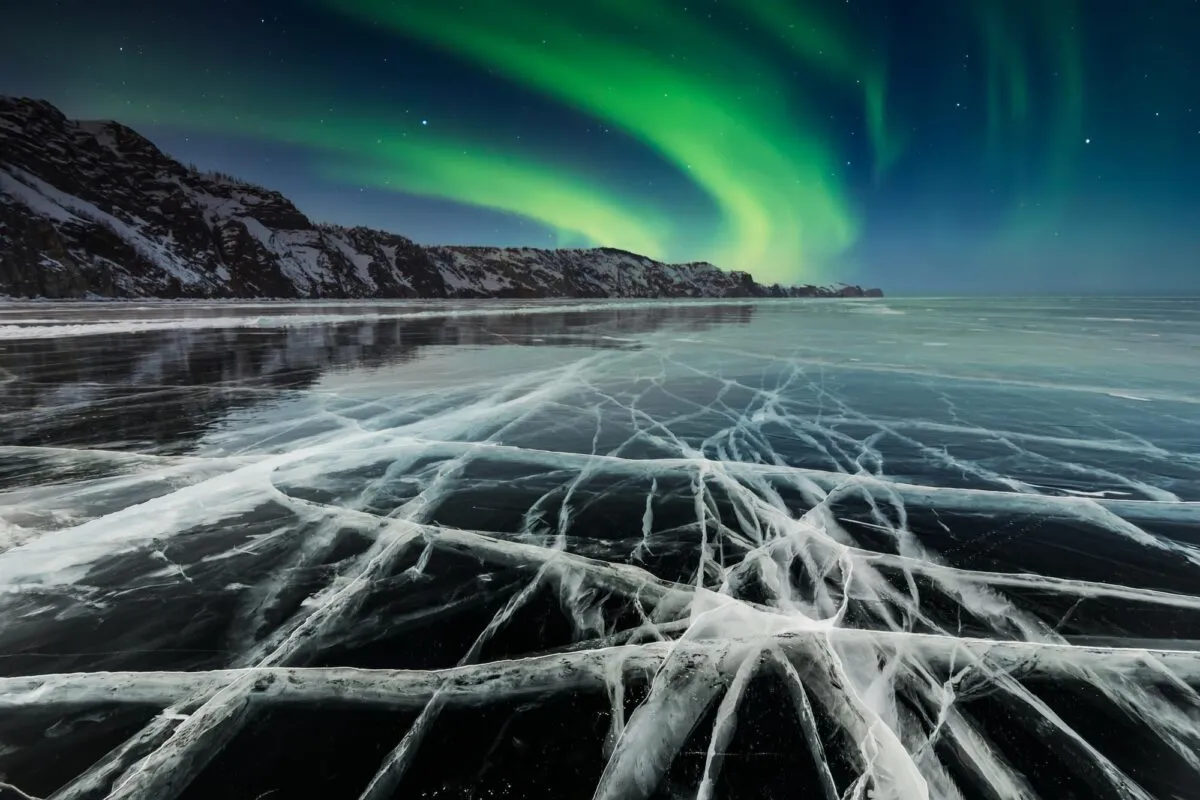 Aurora boreal verde no céu noturno sobre um lago congelado e rachado, com falésias nevadas ao fundo