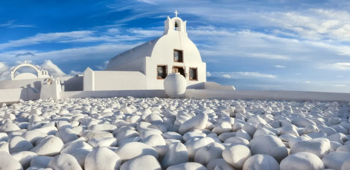 Igreja grega caiada de branco com sino e seixos brancos ao redor, sob céu azul em Santorini