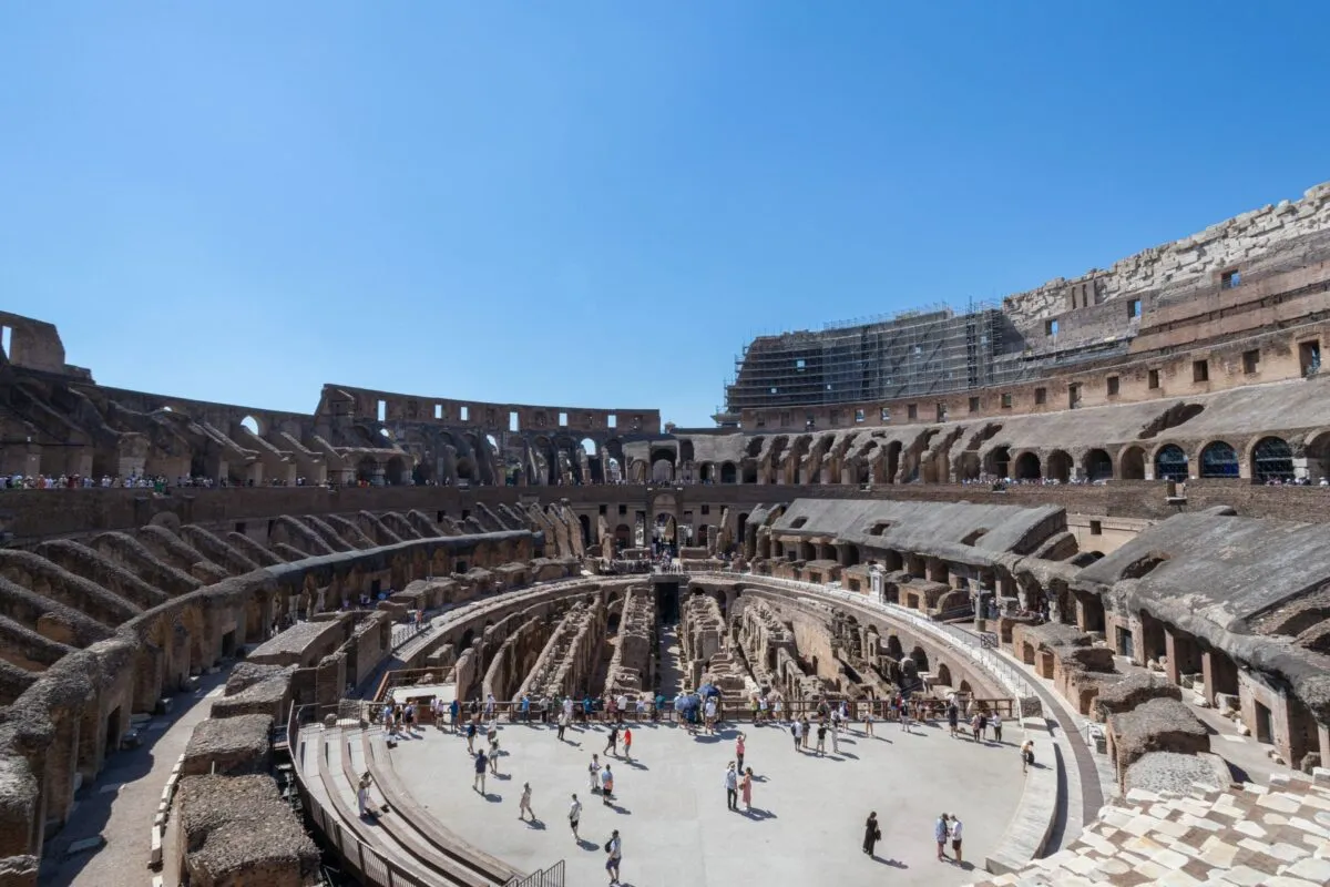 Vista do interior do Coliseu de Roma, na Itália, com turistas na arena e a estrutura de arcos sob céu azul