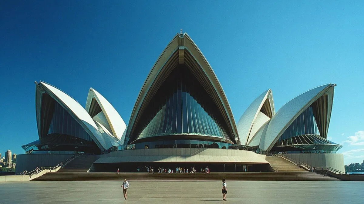 Fachada da Ópera de Sydney com suas conchas brancas sob céu azul, com pessoas caminhando na esplanada em frente ao monumento