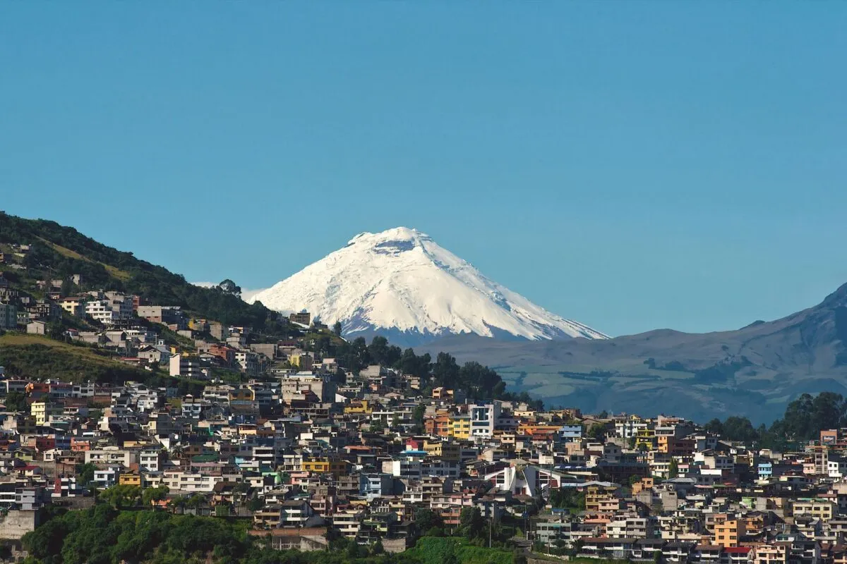 Cidade de Quito espalhada pela encosta de uma colina, com o vulcão Cotopaxi nevado ao fundo sob céu azul
