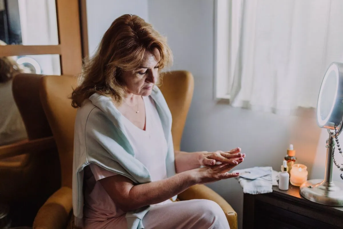 Mulher adulta sentada perto da janela dosa produto de skincare na mão durante rotina de cuidados com a pele madura