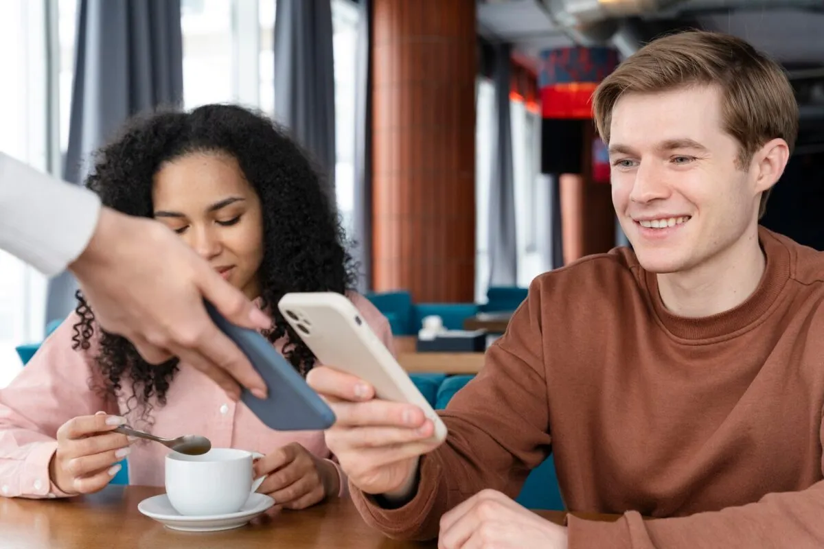 Um homem e uma mulher em um café, usando celulares e sorrindo, com uma xícara de café à frente.
