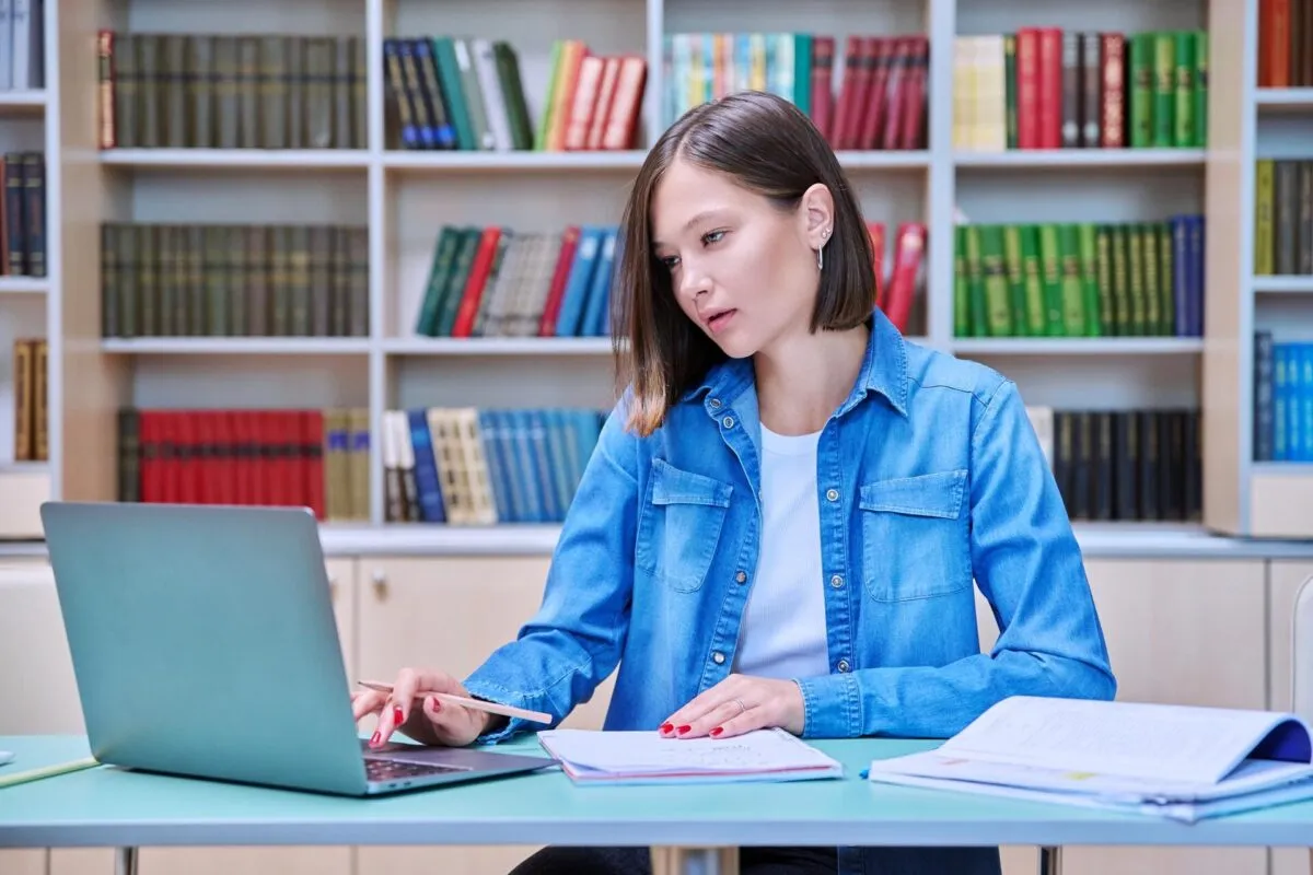 Jovem mulher com camisa jeans estudando com laptop em biblioteca.